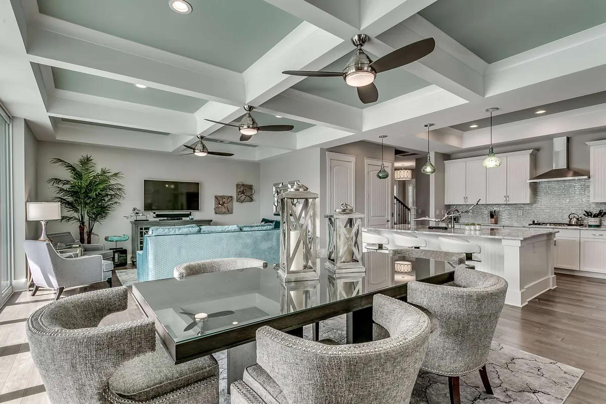 A welcoming dining area featuring a stylish table set for a meal, ceiling fans overhead, lush green plants, and a view into the adjacent kitchen. The setup is ideal for both family dinners and casual brunches, emphasizing the flow between dining and kitchen spaces.