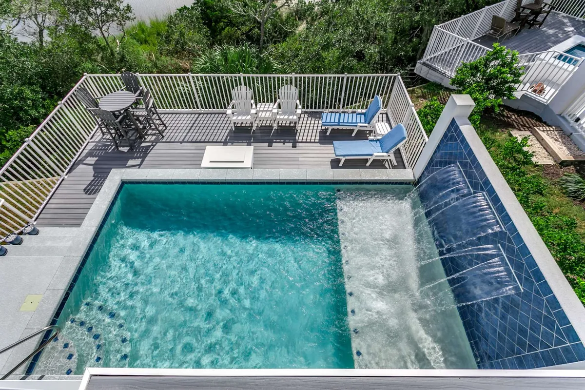 A tranquil private swimming pool with a waterfall feature forms the centerpiece, surrounded by lounge chairs and an outdoor dining set. The pool overlooks a lush, peaceful backdrop, hinting at the nearby saltwater swash. The setup invites both daytime relaxation and lively gatherings.