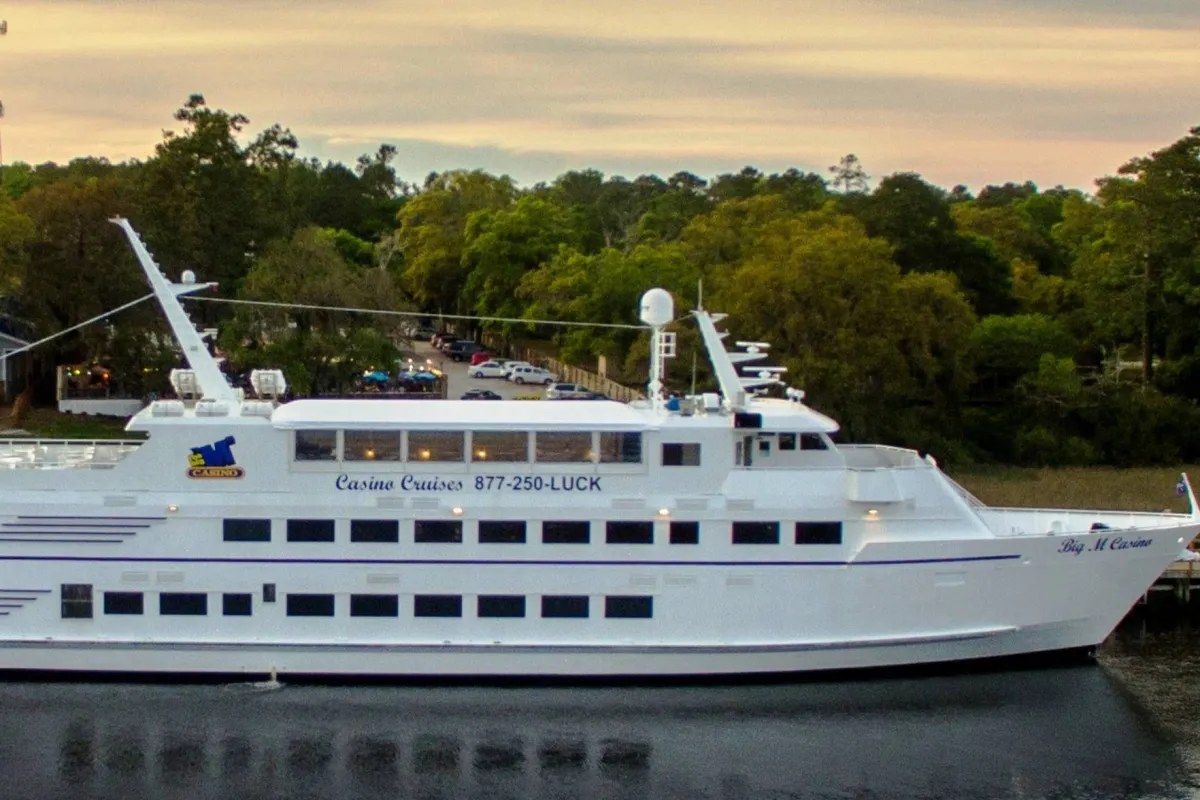 A festive cruise ship, the Barefoot Queen, docked amid lush greenery and illuminated by the setting sun. The scene captures the outdoor excitement of boarding for a riverboat dinner cruise, with the vessel's 'casino' lettering hinting at onboard entertainment and dining options.
