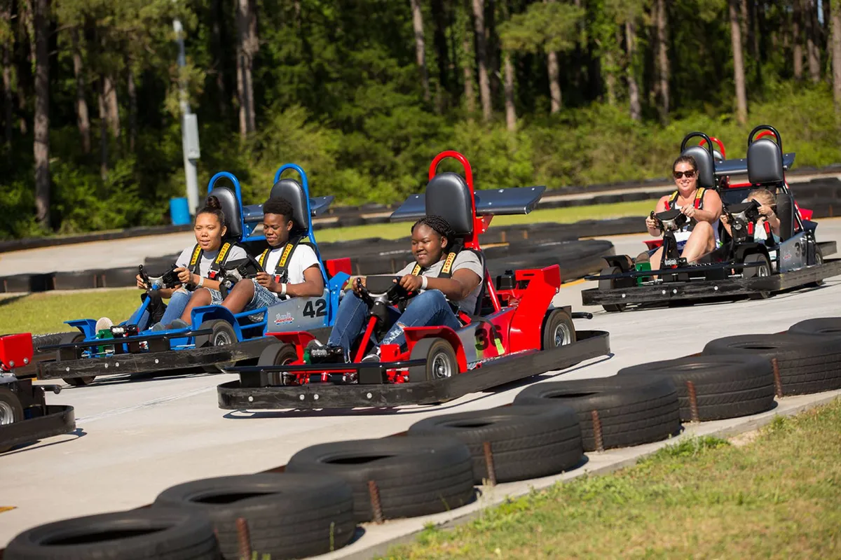 An outdoor go-kart race track featuring participants racing and enjoying themselves, surrounded by trees. The scene is vibrant and full of movement, with families and kids engaging in a thrilling activity together.