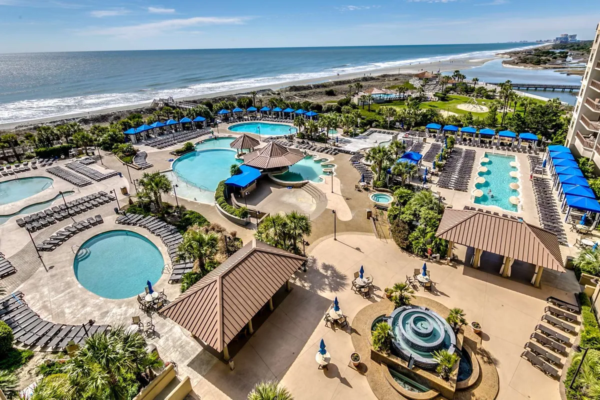A sprawling swimming pool complex set against a beachfront backdrop, dotted with lounge chairs, umbrellas, and palm trees. The atmosphere is lively yet relaxing, with guests enjoying the sun, water, and nearby fire pits as evening approaches.