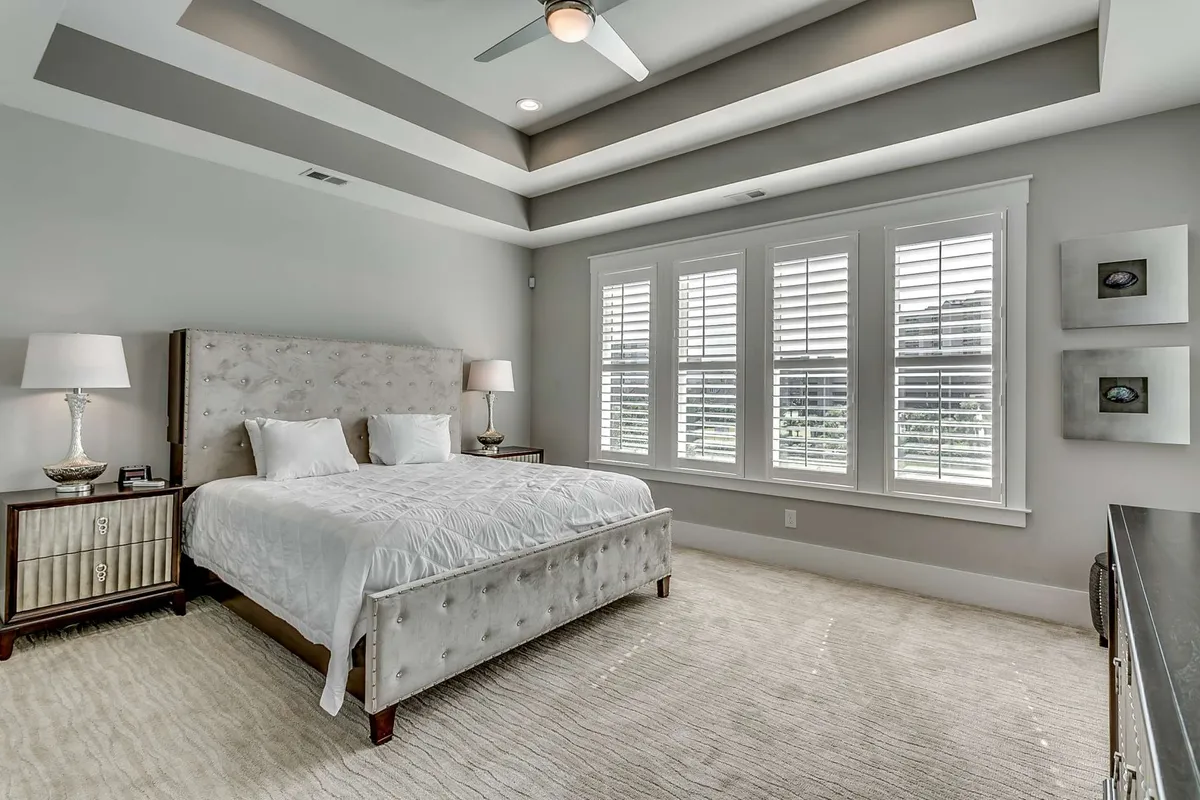 A serene bedroom scene featuring a comfortable bed placed against a wall with a large window letting in soft natural light. The room includes matching nightstands, lamps, and tasteful artwork, conveying a sense of tranquility and comfort.