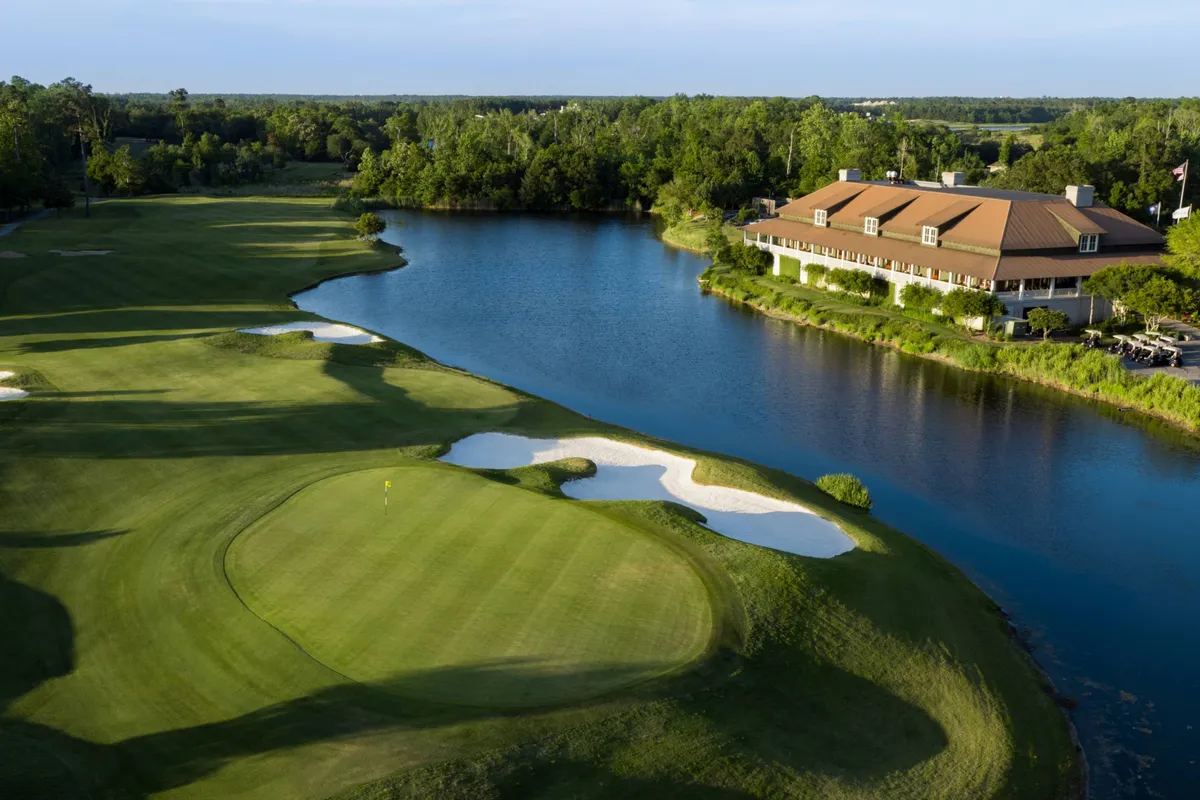 A sweeping view of a lush golf course bordered by a tranquil lake, with the clubhouse visible in the distance and sand traps dotting the greens. The setting is serene and inviting, perfect for a relaxing round of golf or social gathering.