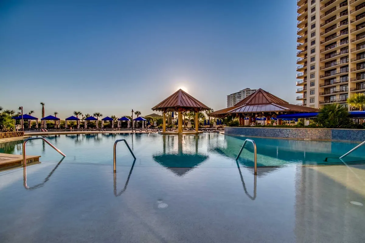 An infinity pool surrounded by thatched-roof cabanas, plush lounge chairs, and palm trees, with a modern high-rise building in the background. The setting exudes tranquility and exclusivity, ideal for unwinding or socializing at the water's edge.