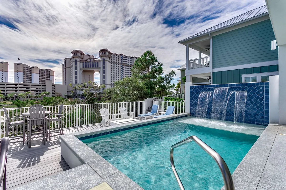 The image showcases the luxurious exterior of a single-family home in North Myrtle Beach, centered around a sparkling swimming pool with an elegant waterfall feature. Plush lounge chairs line the poolside, inviting relaxation, while lush greenery creates a private oasis. In the background, the warm glow of sunset casts a tranquil light, with distant buildings hinting at a peaceful yet vibrant location.