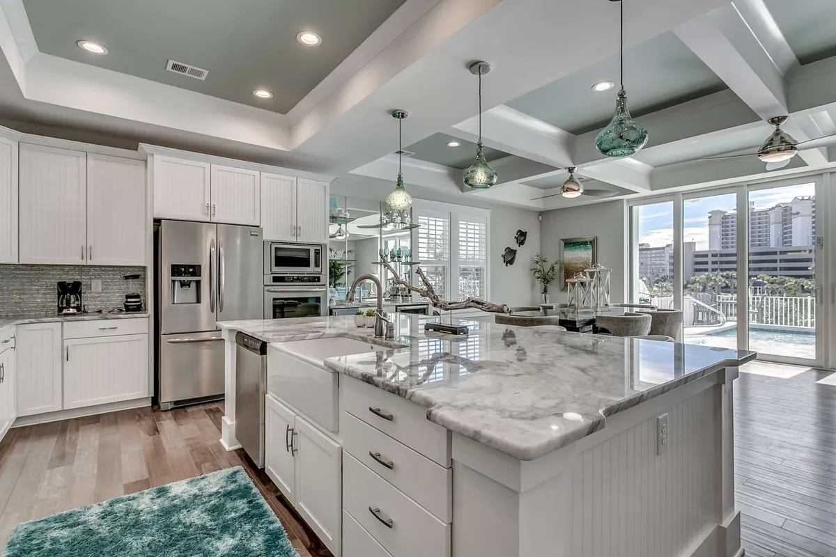 A spacious, sunlit open area that blends a modern kitchen with a large island, marble countertops, and stylish hanging pendant lights. Expansive windows line the space, offering inviting views of the shimmering pool beyond. The scene is set up for lively cooking, entertaining, and group gatherings, giving a sense of warmth and liveliness.