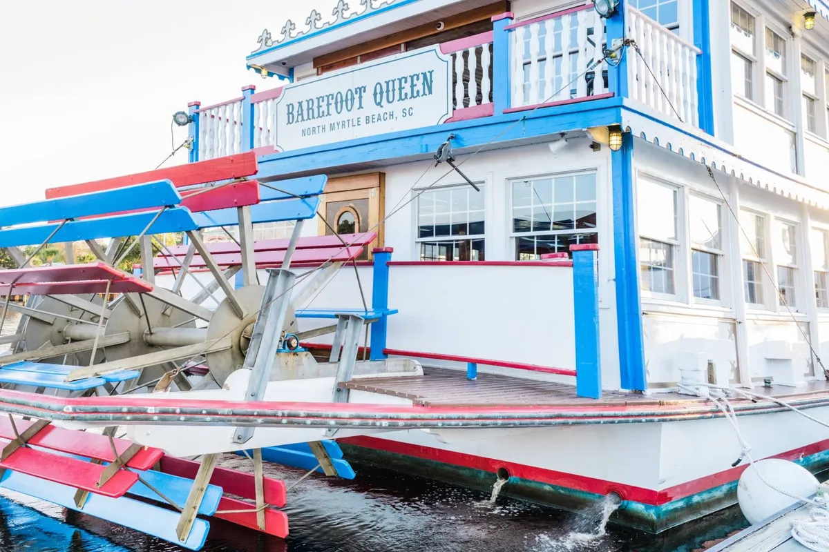 A lively exterior shot of a vibrant paddlewheel boat with a colorful facade and inviting signage. The scene hints at festive river cruises, waterside dining, and social celebrations at sunset.