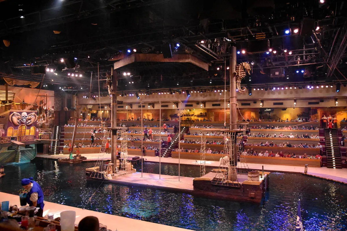 A vibrant interior setting of a theater with a stage featuring aquatic elements, decorative ship, and guests seated in anticipation. The space is dynamic, hinting at immersive performances and interactive entertainment experiences.